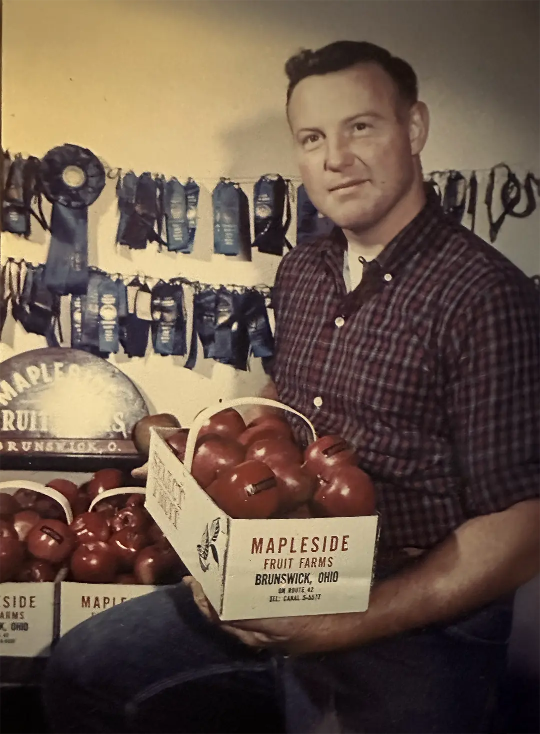 A young Apple Bill Eyssen holding a Mapleside Fruit Farms crate of apples, prize ribbons behind him